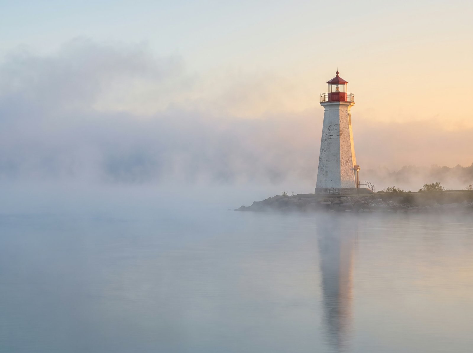 A white lighthouse on a calm shoreline in soft morning mist